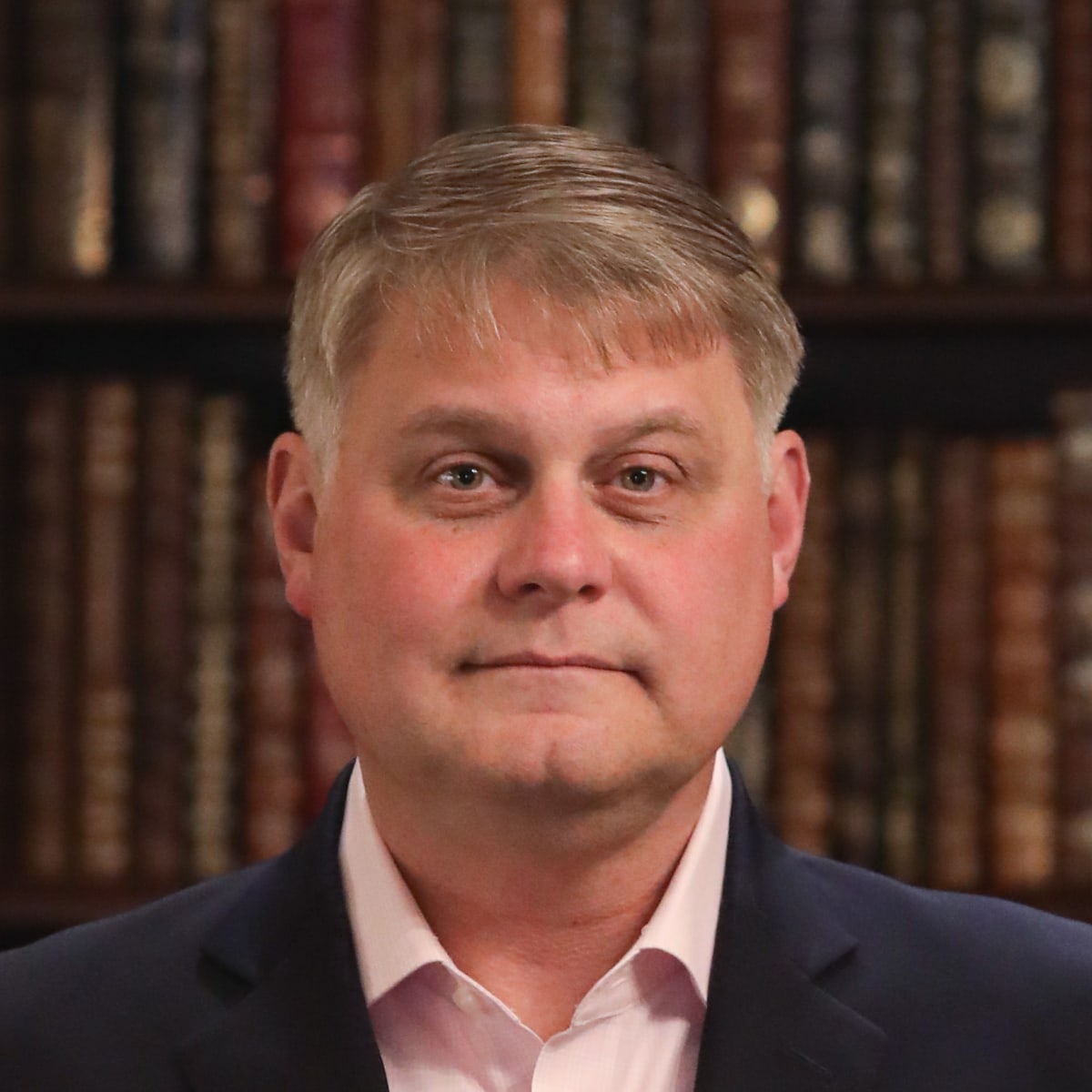 Christopher Nuneviller; smiling middle-aged man with clean face and button up shirt with suit jacket in front of bookcase